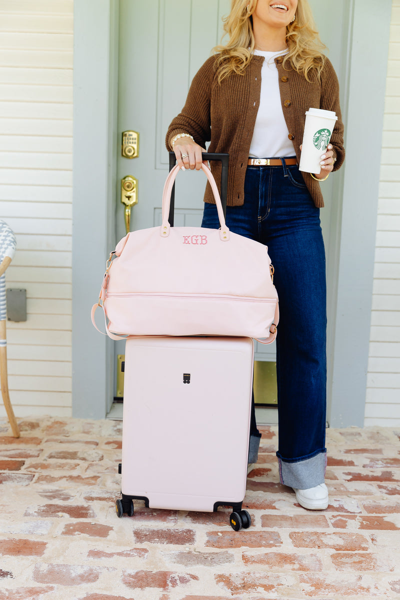 A woman stands on a brick porch in front of a light green door, holding a pink hard-shell suitcase and stacking a matching pink duffel bag on top. She wears a brown cardigan, white t-shirt, and blue jeans, and holds a Starbucks coffee cup.