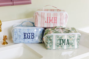 Three personalized toiletry bags in different patterns and colors are arranged on a white bathroom counter next to a gold faucet. The top bag is pink and white striped with the initials 'JMH' in pink.