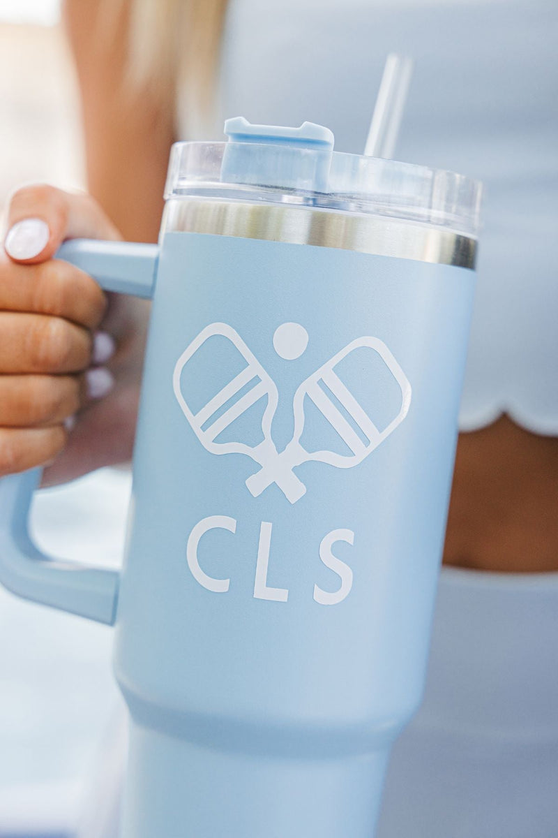 A girl in a blue outfit holds up a blue 40 oz tumbler with a white pickleball and monogram design