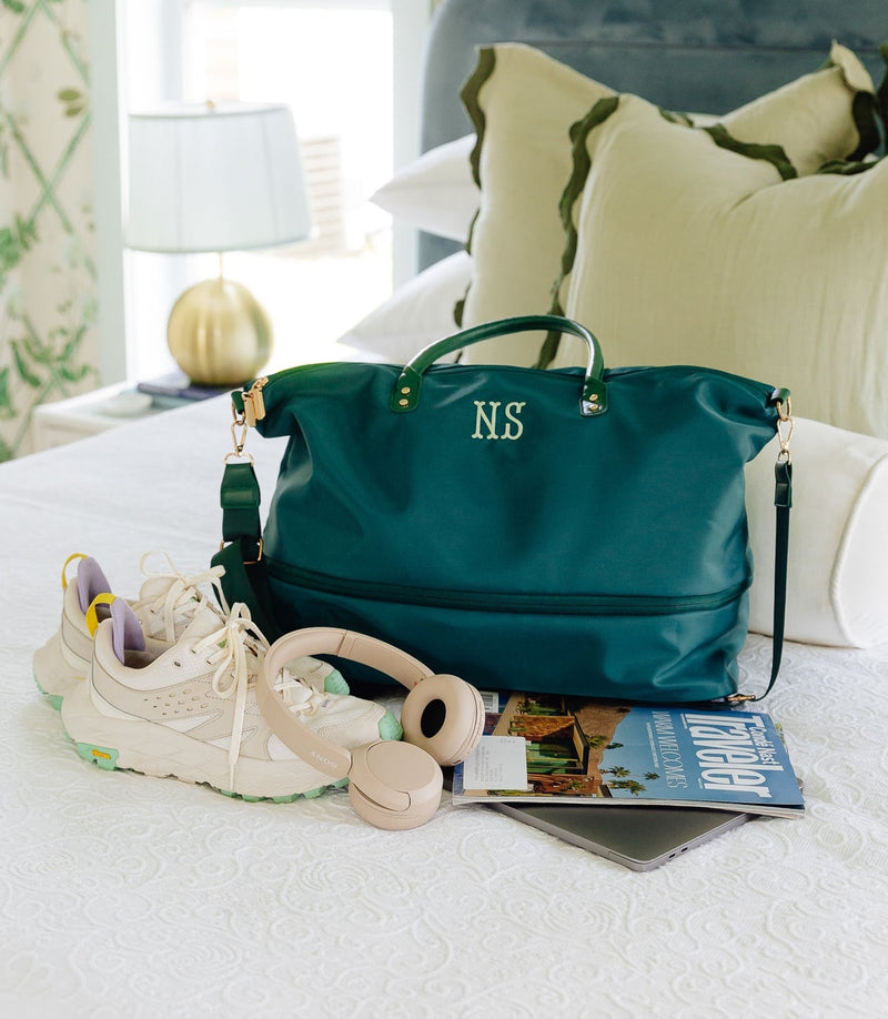 A teal leather monogram duffel bag with gold hardware sits on a white bed, styled with a pair of cream sneakers, a beige cordless phone, a magazine, and a gray tablet. The background features green and white floral wallpaper and a blue-gray headboard.