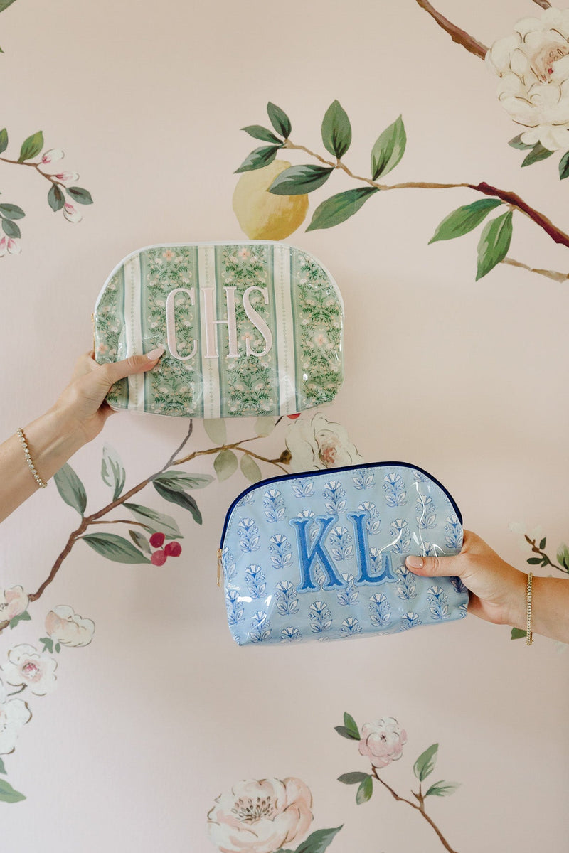 Two personalized cosmetic pouches are held by hands against a floral wallpaper background. The top pouch is green and white striped with the monogram 'ICS' in white.