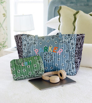 A blue and navy patterned tote bag with the name 'PARKER' embroidered in colorful letters sits on a white bed, alongside a matching green patterned pouch, wooden headphones, and a tablet. The background shows a softly lit bedroom with patterned curtains and pillows.