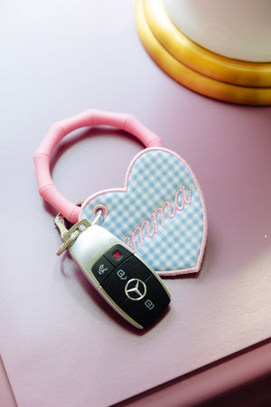 A pink silicone keychain with a heart-shaped bag tag featuring a light blue and white gingham pattern and the embroidered name 'Emma', holding a Mercedes-Benz key fob, displayed on a pink night stand.