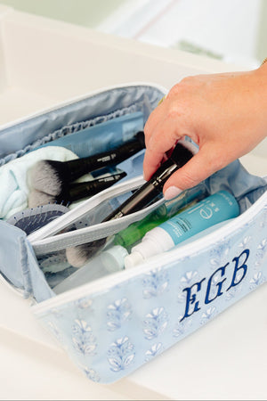 A light blue floral makeup bag with the monogram 'EGB' on the front, shown open with cosmetics inside. A person's hand is reaching into the bag, which is placed on a white bathroom counter.