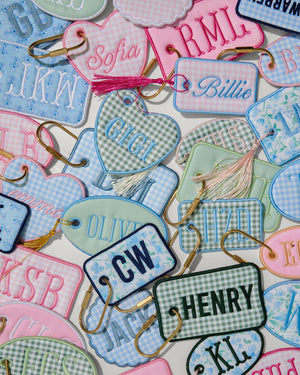 A dense flat-lay collection of personalized embroidered name tags in pink, light blue, mint green, and white gingham patterns, each with a gold metal clip and a small tassel.