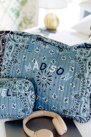 A blue paisley-patterned quilted gift set including a large bag, a smaller pouch, and a tablet sleeve, with custom monogram letters 'MADISON' and 'ELI', styled with beige headphones on a laptop in a bright room.