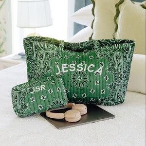 A green and white paisley-patterned tote bag with the name 'JESSICA' printed in white on the front, displayed on a bed in a bedroom setting next to a matching pouch.