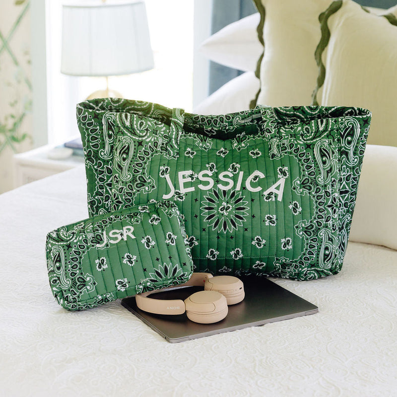 A green and white paisley-patterned tote bag with the name 'JESSICA' printed in white on the front, displayed on a bed in a bedroom setting next to a matching pouch.