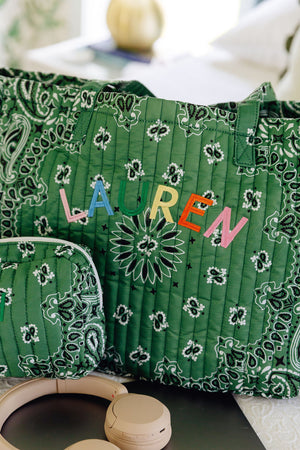A green quilted tote bag with a white paisley bandana pattern, featuring the name 'LAUREN' embroidered in colorful letters across the front. A matching zippered pouch is partially visible on the left, and a pair of beige headphones rests at the bottom of the frame.
