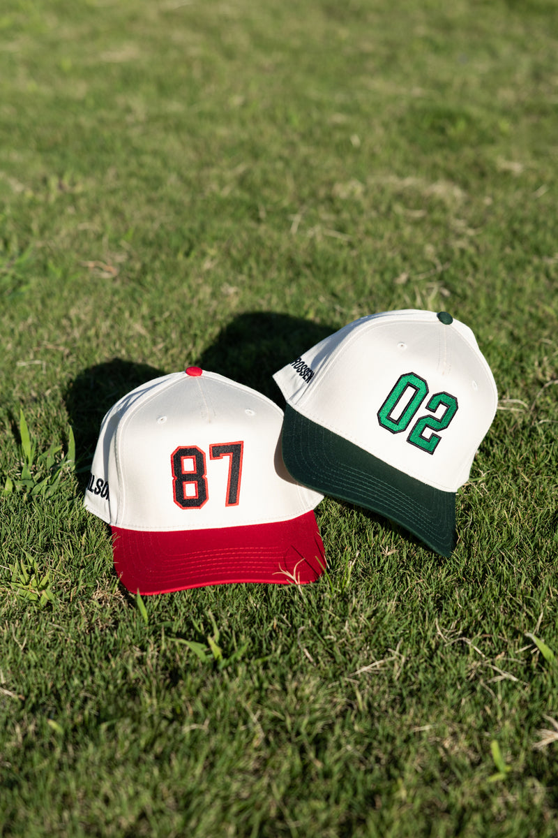 Field close-up: red-brim “87” and green-brim “02” Embroidered Jersey Number Vintage Trucker Hats.