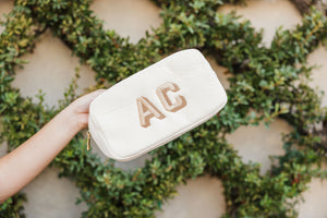 A person holds up a white corduroy pouch with custom initials against a wall with leaves.
