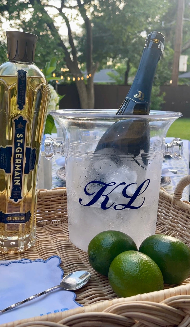 Clear acrylic ice bucket monogrammed “KL” in navy, styled outdoors with chilled bottle, fresh limes, a St-Germain bottle, and scalloped “JMD” cocktail napkin.