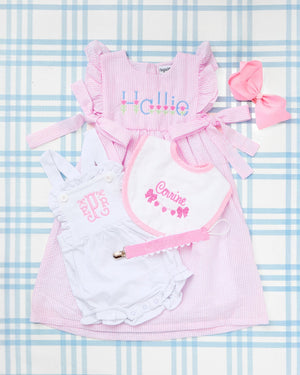Flat lay of charming baby girl essentials displayed on a blue gingham background, featuring the standout Seersucker Bow Dress in pink with ruffle sleeves, side bows, and an embroidered tulip motif with the name “Hallie.” Paired with a white Cotton Sunsuit showcasing elegant pink monogramming, a coordinating embroidered bib with the name “Corinne” and bow motif, a pink pacifier clip, and a matching pink hair bow—this personalized set is perfect for springtime gifting or stylish everyday wear.