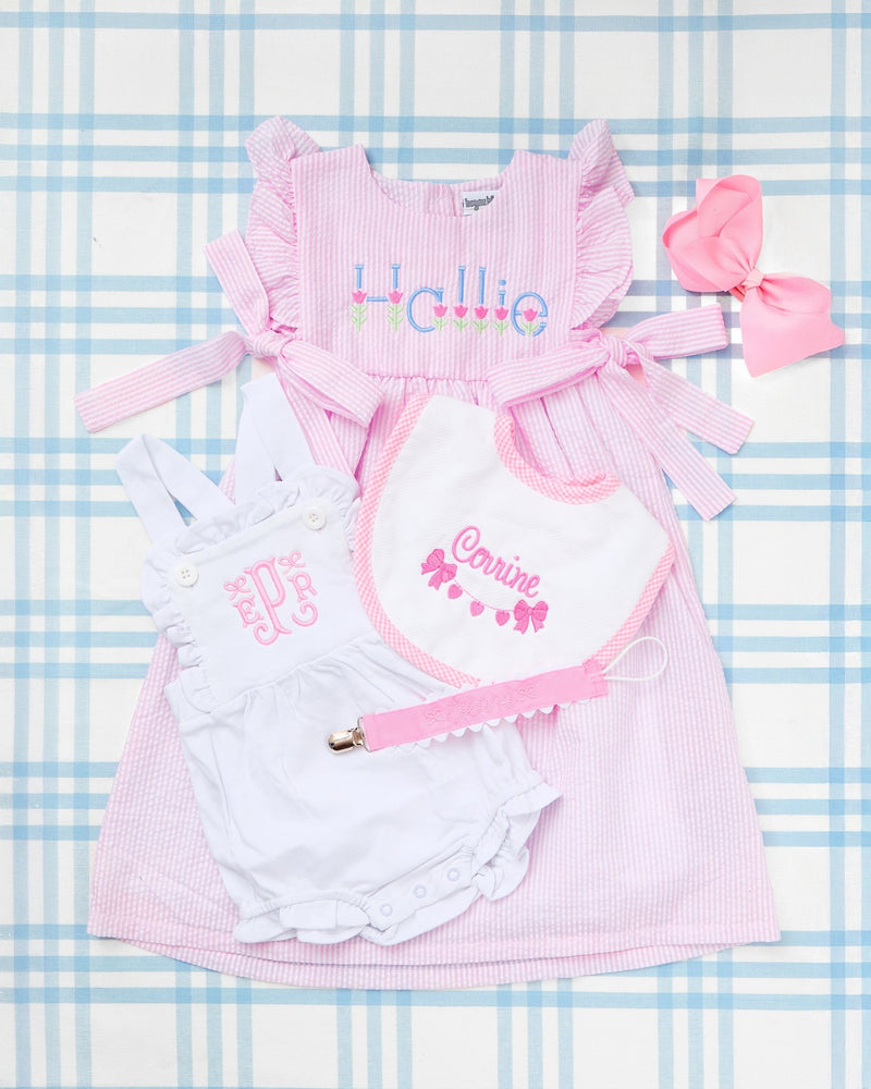 Flat lay of charming baby girl essentials displayed on a blue gingham background, featuring the standout Seersucker Bow Dress in pink with ruffle sleeves, side bows, and an embroidered tulip motif with the name “Hallie.” Paired with a white Cotton Sunsuit showcasing elegant pink monogramming, a coordinating embroidered bib with the name “Corinne” and bow motif, a pink pacifier clip, and a matching pink hair bow—this personalized set is perfect for springtime gifting or stylish everyday wear.