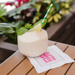 A cocktail napkin with a pink embroidered "cheers" design is placed under a drink in a coconut cup.