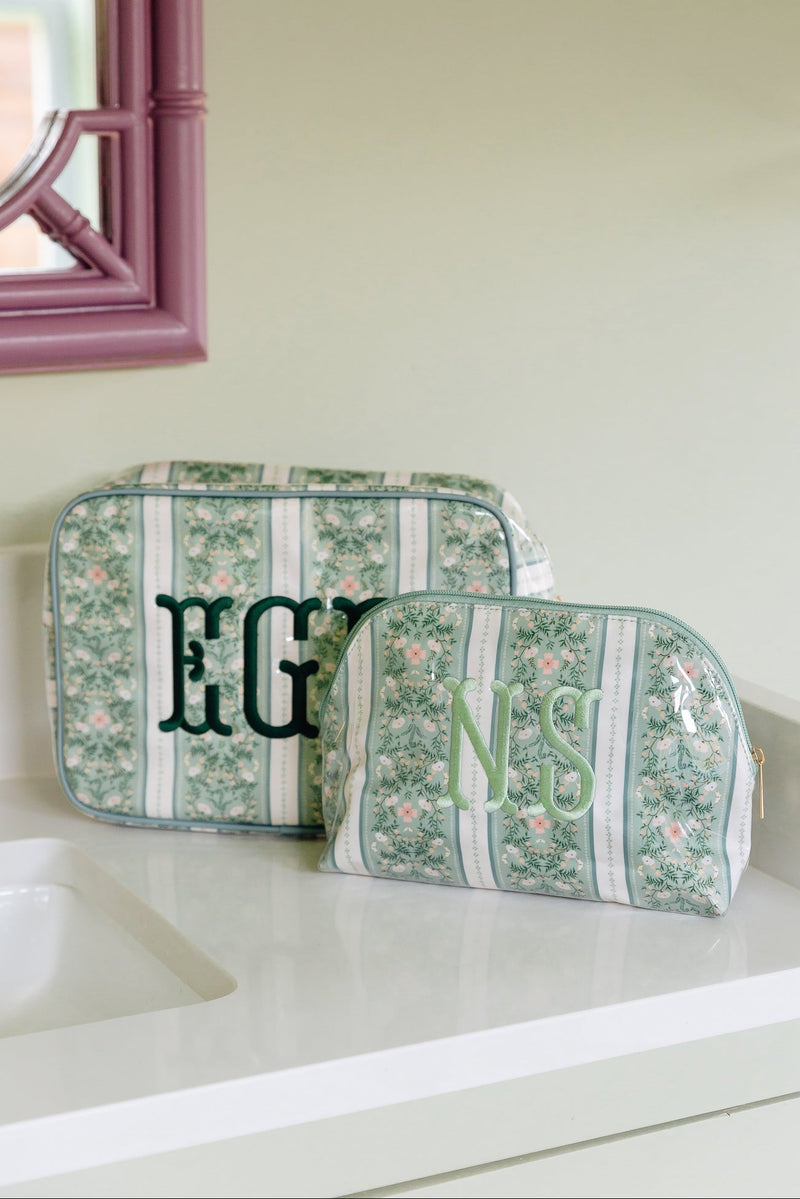 Sage green floral clear coated pouch monogrammed 'EGB' in dark green and a smaller matching pouch monogrammed 'NS' in light green, on a bathroom counter.