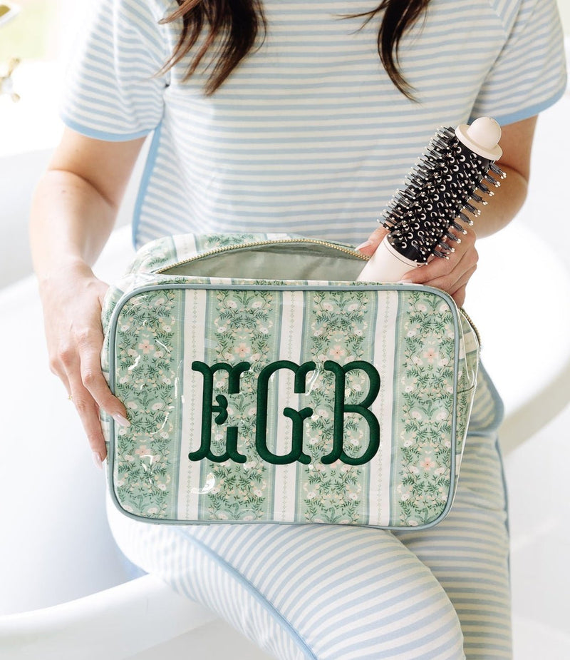 A woman in a bathroom setting wearing a light blue and white striped pajama set, holding a green and white floral toiletry bag with the letters 'EGB' monogrammed on the front, while holding a black hairbrush.