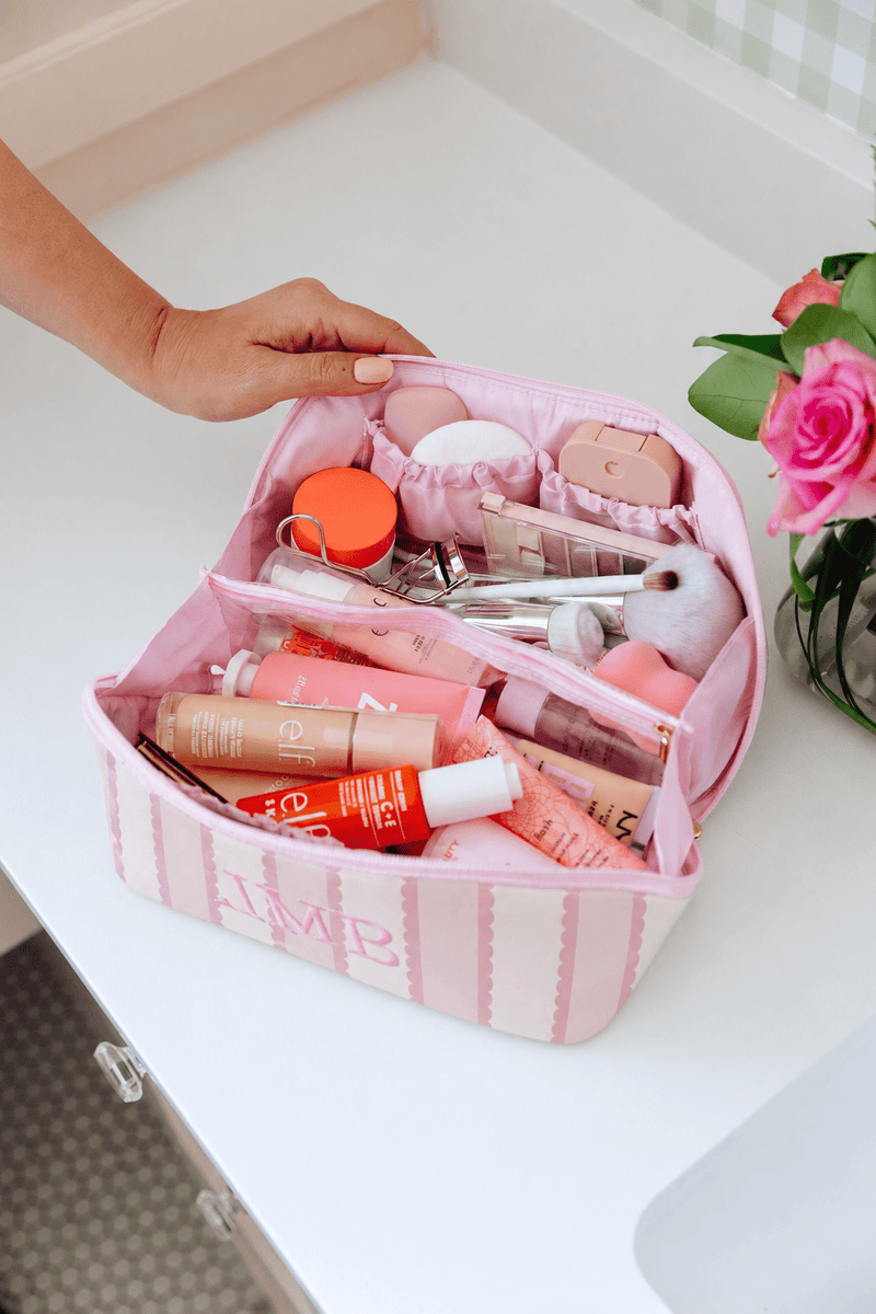 A pink striped cosmetic bag with a monogrammed 'JMR' logo, filled with various makeup items, held open by a hand on a white bathroom counter next to pink roses.