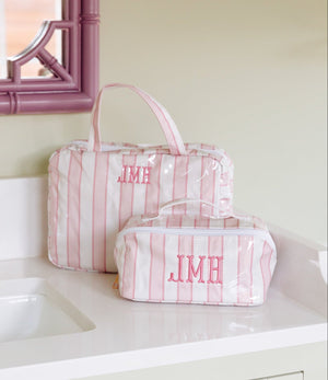 Two pink and white striped toiletry bags with pink monogrammed initials 'JMH' sit on a white bathroom counter next to a sink and a pink-framed mirror.
