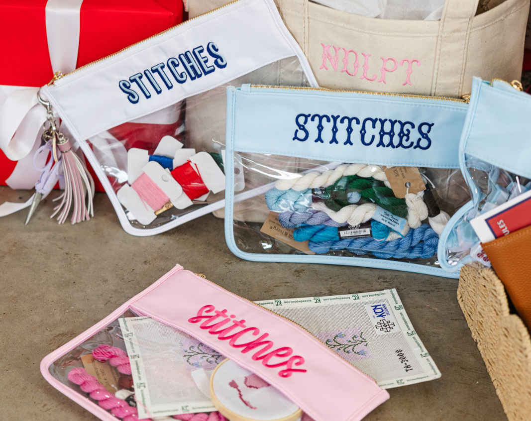 Needlepoint gift collection with monogrammed tumblers, pouches, and blankets arranged by a tree.