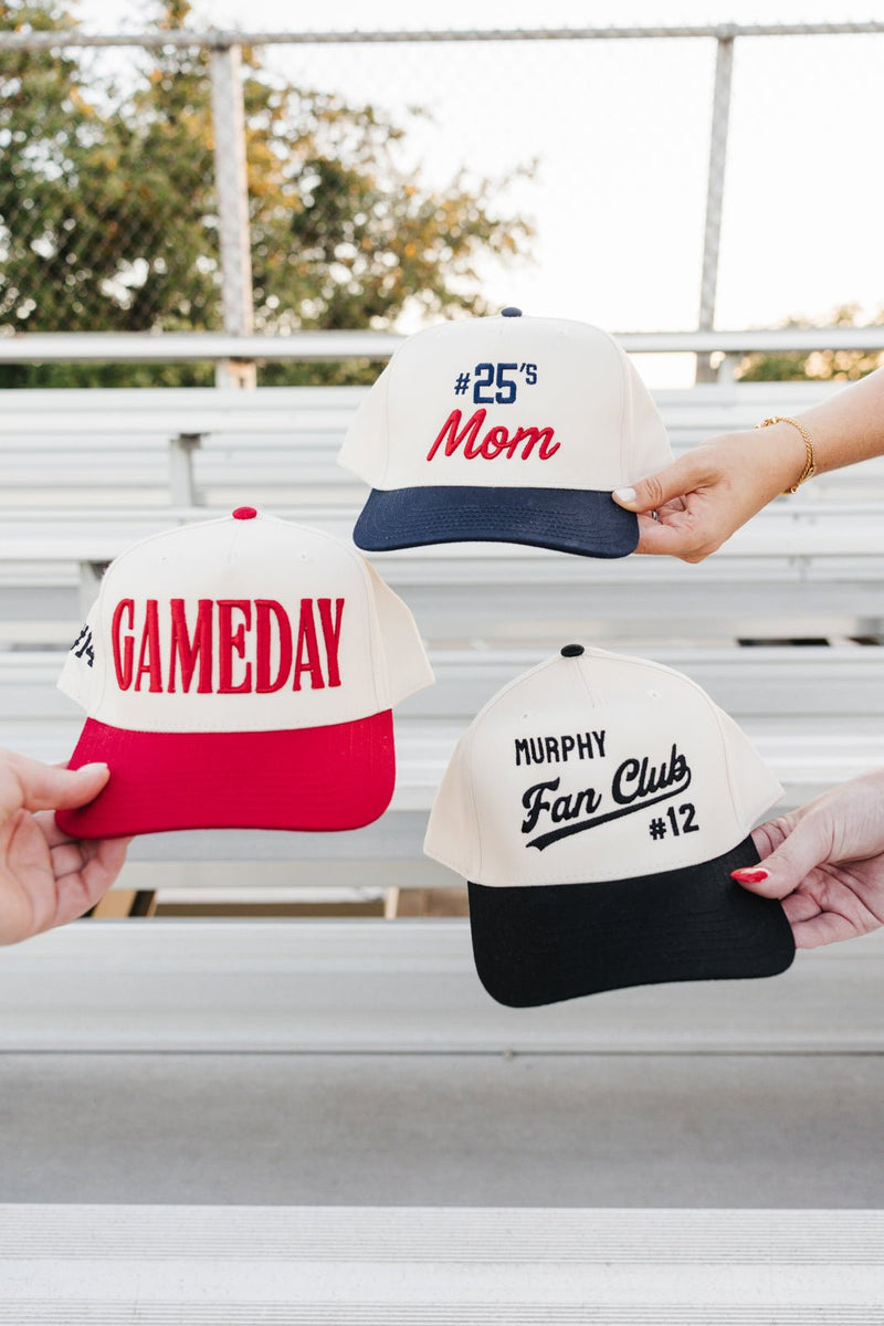 Hands display Custom #'s Mom 3D Puff Vintage Trucker Hat beside Gameday and Fan Club caps on bleachers.