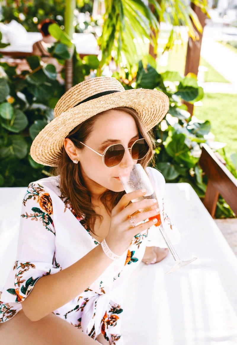 Woman relaxing outdoors in tropical surroundings, sipping a drink while wearing a Custom Boater Hat with a black band, round sunglasses, and a floral wrap dress—capturing effortless resort style and laid-back luxury.