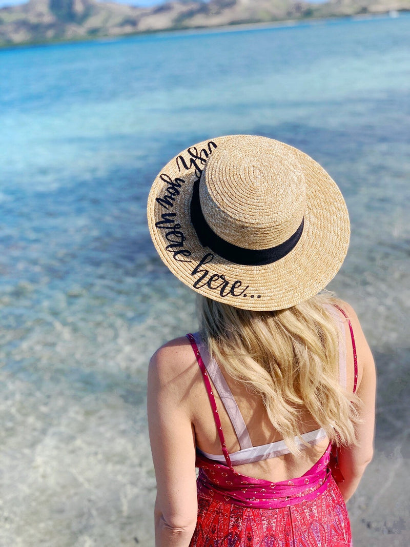 Woman standing in shallow turquoise water wearing a wide-brimmed custom boater hat with black ribbon and playful script that reads “blah blah blah… here.” The straw hat adds a chic, personalized touch to her pink patterned sundress, perfect for a beachside escape.