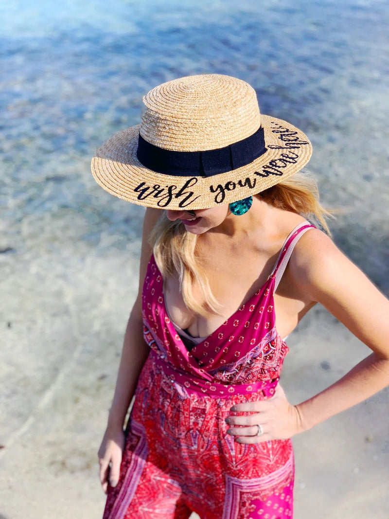 Woman standing by the shoreline wearing a wide-brimmed Custom Boater Hat with a black ribbon and playful embroidered script that reads “wish you were here,” styled with a vibrant red printed dress and turquoise statement earrings—perfect for beach days and vacation vibes.
