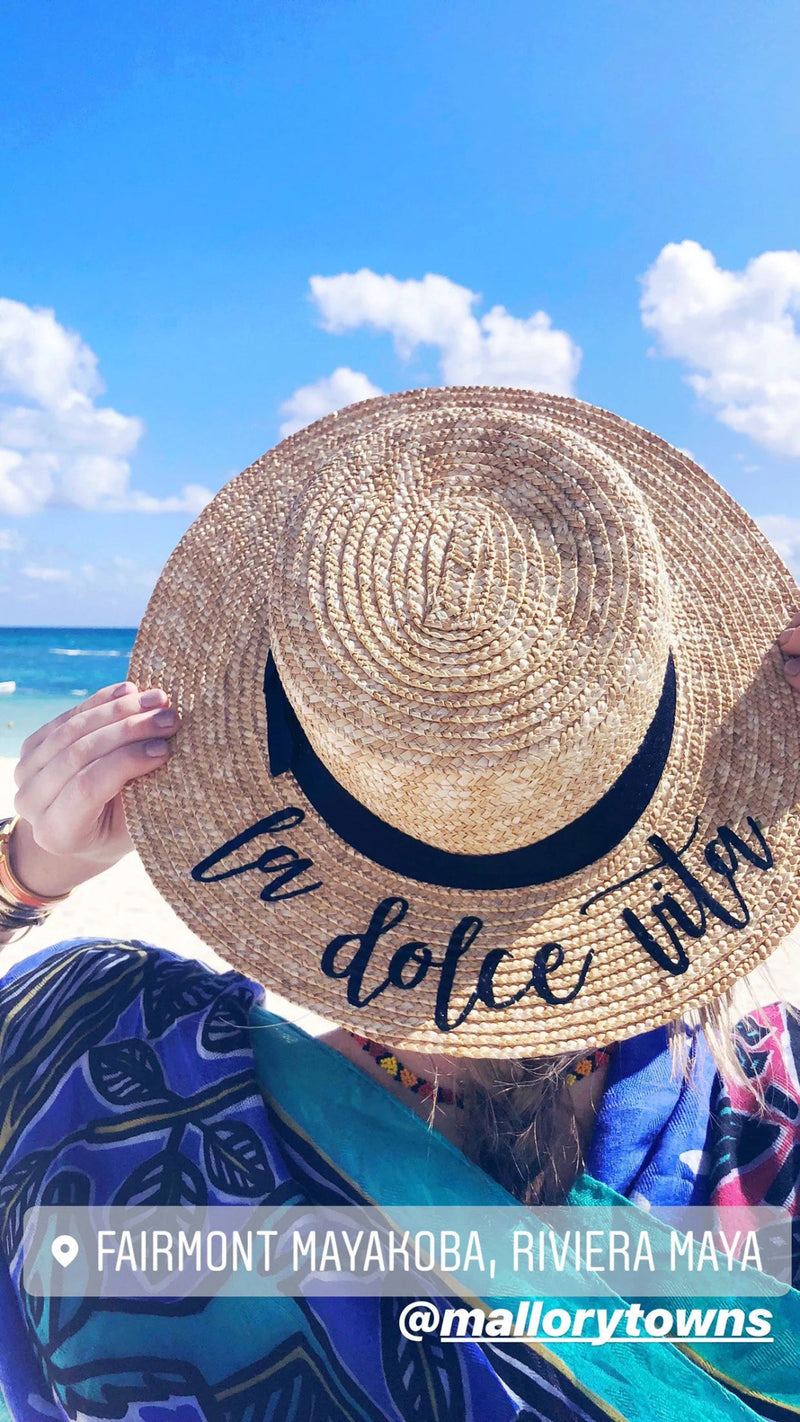 Woman holding up a Custom Boater Hat embroidered with “la dolce vita” against a vibrant beach backdrop at Fairmont Mayakoba, Riviera Maya—perfectly capturing a carefree coastal escape and personalized travel style.
