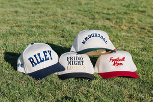 Cream trucker hats on grass; center shows “FRIDAY NIGHT LIGHTS” in black and gold—Embroidered Friday Night Lights Vintage Hat.