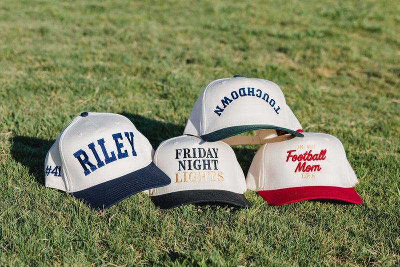 Cream trucker hats on grass; center shows “FRIDAY NIGHT LIGHTS” in black and gold—Embroidered Friday Night Lights Vintage Hat.