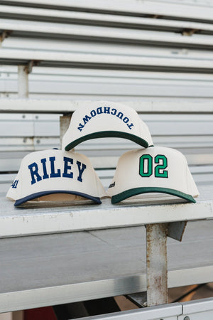 Custom Collegiate Vintage Trucker Hats on bleachers: cream caps with navy “RILEY”, green “02”, and arched “TOUCHDOWN”.