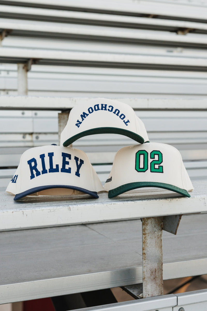 Custom Collegiate Vintage Trucker Hats on bleachers: cream caps with navy “RILEY”, green “02”, and arched “TOUCHDOWN”.