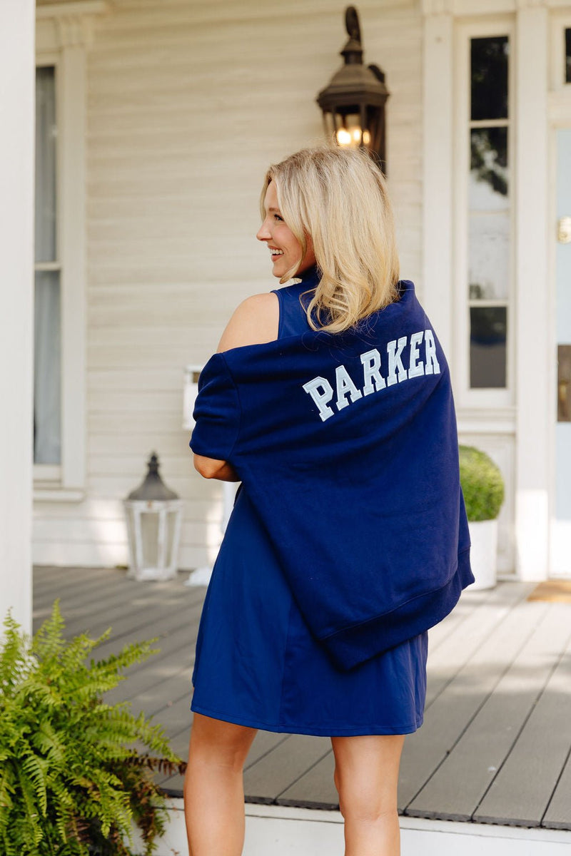 Woman standing on a porch, looking over her shoulder wearing an off-shoulder navy mockneck sweatshirt printed 'PARKER' on the back