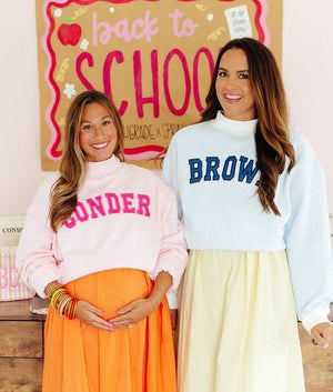 Two women standing side-by-side in matching light-colored sweatshirts with colorful text. The woman on the left wears a light pink sweatshirt with 'GONDER' in pink, paired with an orange skirt.
