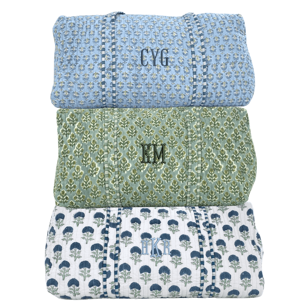 Stack of embroidered block print duffel bags in blue, green, and white floral prints with monogram personalization.
