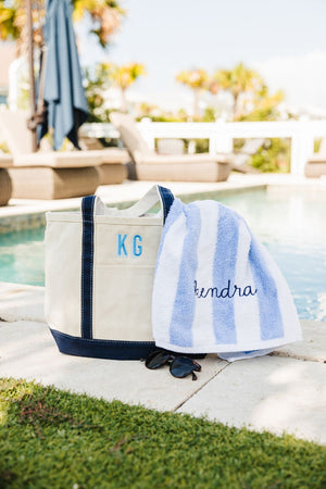 A navy canvas tote is placed next to a pool and is filled with a blue cabana towel with an embroidered name in navy thread.