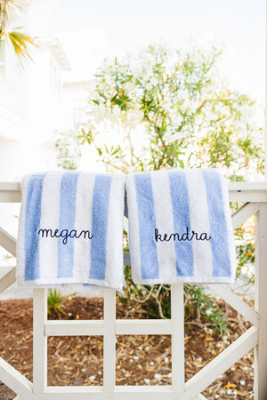 Two blue cabana towels are placed on a railing and are embroidered with names in navy thread.