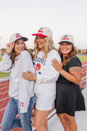 Three women wearing matching 'Mom' hats and personalized sweatshirts on a track field.