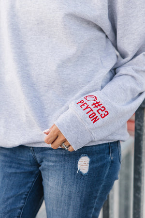Gray sweatshirt with red personalized text on sleeve worn by a person, standing against a neutral background.