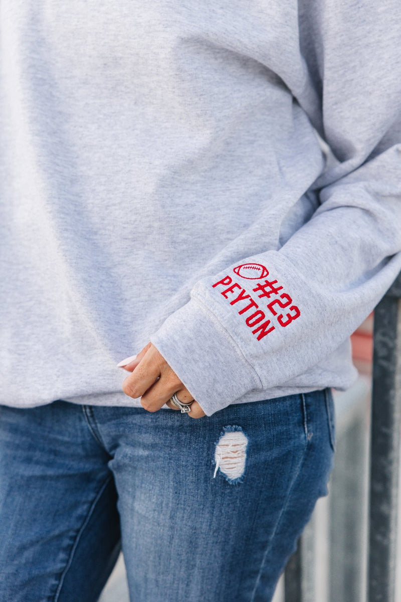 Gray sweatshirt with red personalized text on sleeve worn by a person, standing against a neutral background.