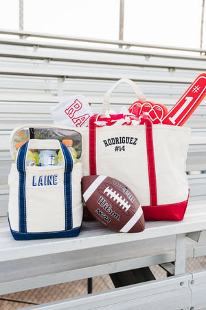 Cream canvas gameday tote with red trim and “Rodriguez #14” embroidery beside a navy-trimmed “Laine” cooler tote and football.