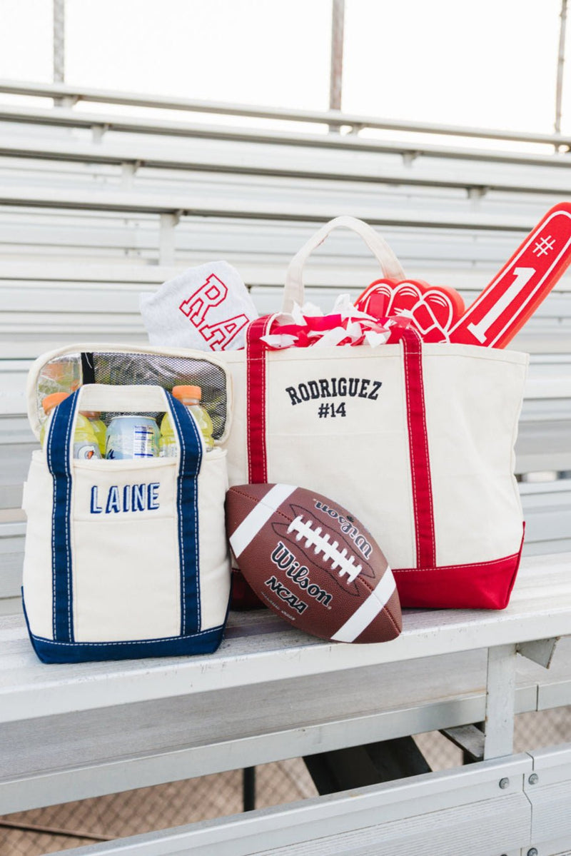Cream canvas gameday tote with red trim and “Rodriguez #14” embroidery beside a navy-trimmed “Laine” cooler tote and football.