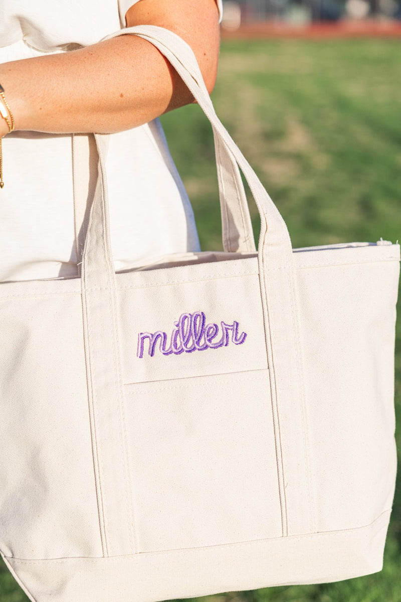 Natural canvas tote with embroidered purple “miller” script across front pocket, carried over shoulder.