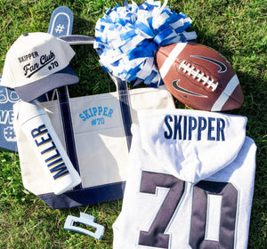 Canvas gameday tote with navy trim and “Skipper #70” embroidery, styled with matching hat, bottle, pom, and football.