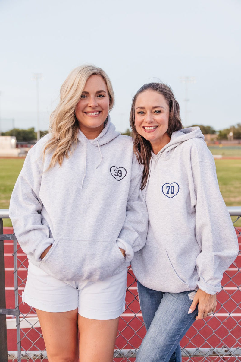 Two women in gray hoodies with navy heart “39” and “70” appliques on chests at field