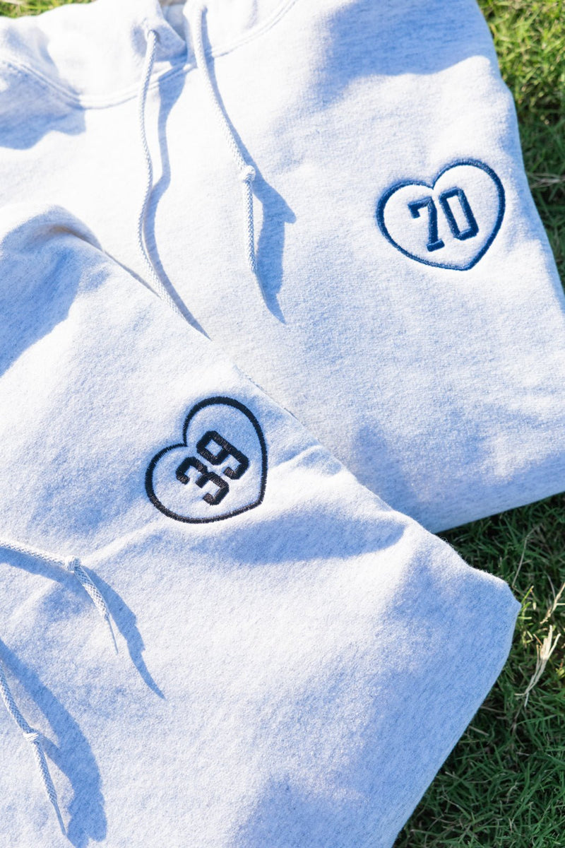 Two gray hoodies on grass each with embroidered heart appliqué numbers 39 and 70 – Embroidered Gameday Hoodie with Applique