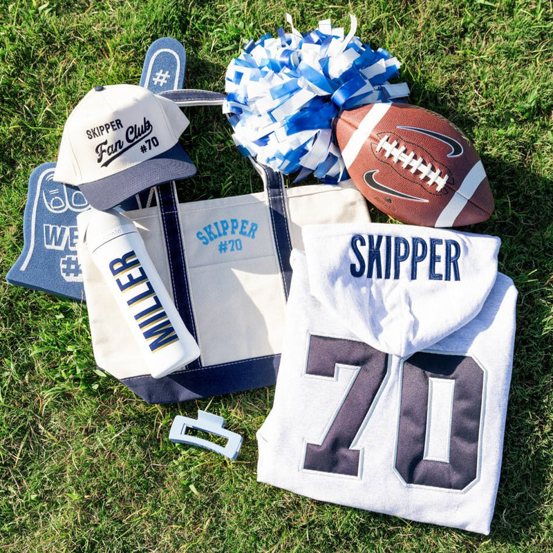 Flat lay of gray hoodie with navy “70,” navy-trim tote, fan club hat, custom water bottle, pompom, and foam finger.
