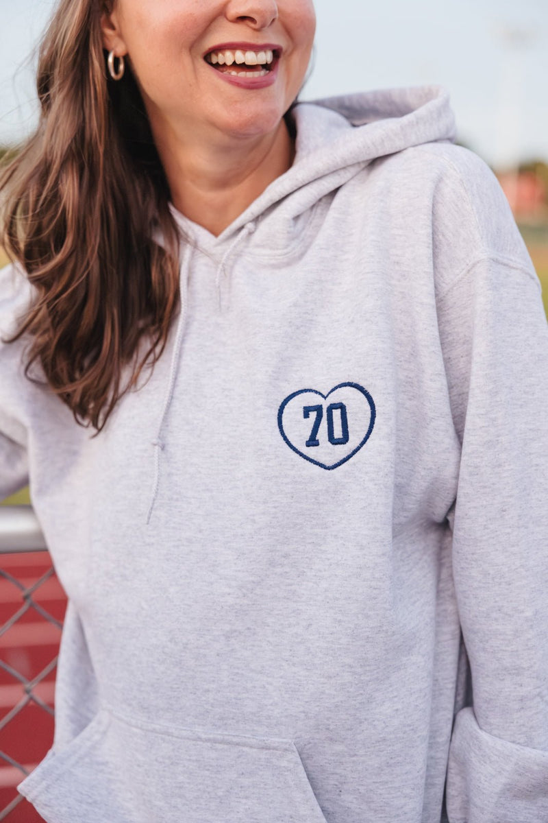 Smiling woman wearing gray hoodie with navy heart-shaped “70” applique on left chest – Embroidered Gameday Hoodie