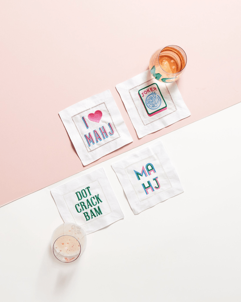 Set of four embroidered cocktail napkins with playful Mahjong-inspired designs and sayings like "I ❤️ MAHJ" and "DOT CRACK BAM," displayed alongside cocktails for a fun, game-night vibe.